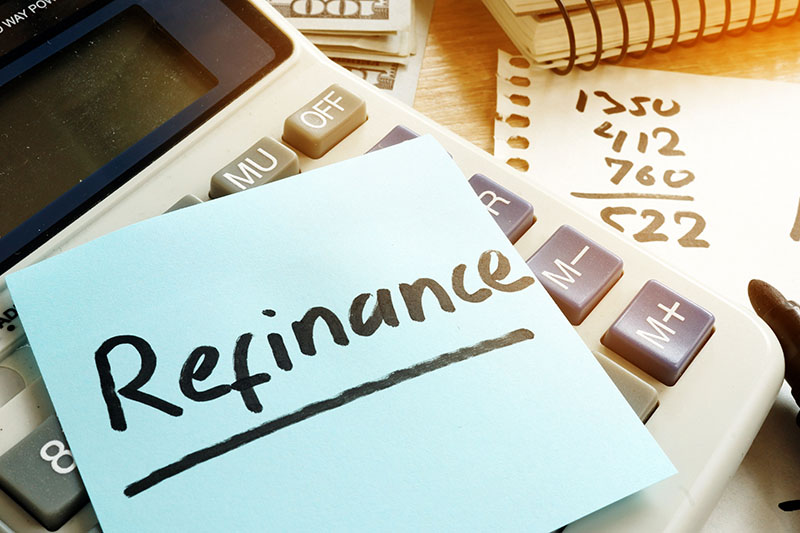 A sticky note with the word Refinance is placed on a calculator. In the background, there are scattered papers with handwritten numbers and a spiral notebook.
