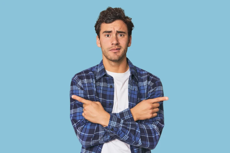 A man in a blue plaid shirt stands against a light blue background, crossing his arms and pointing in opposite directions with both index fingers, looking confused or uncertain.